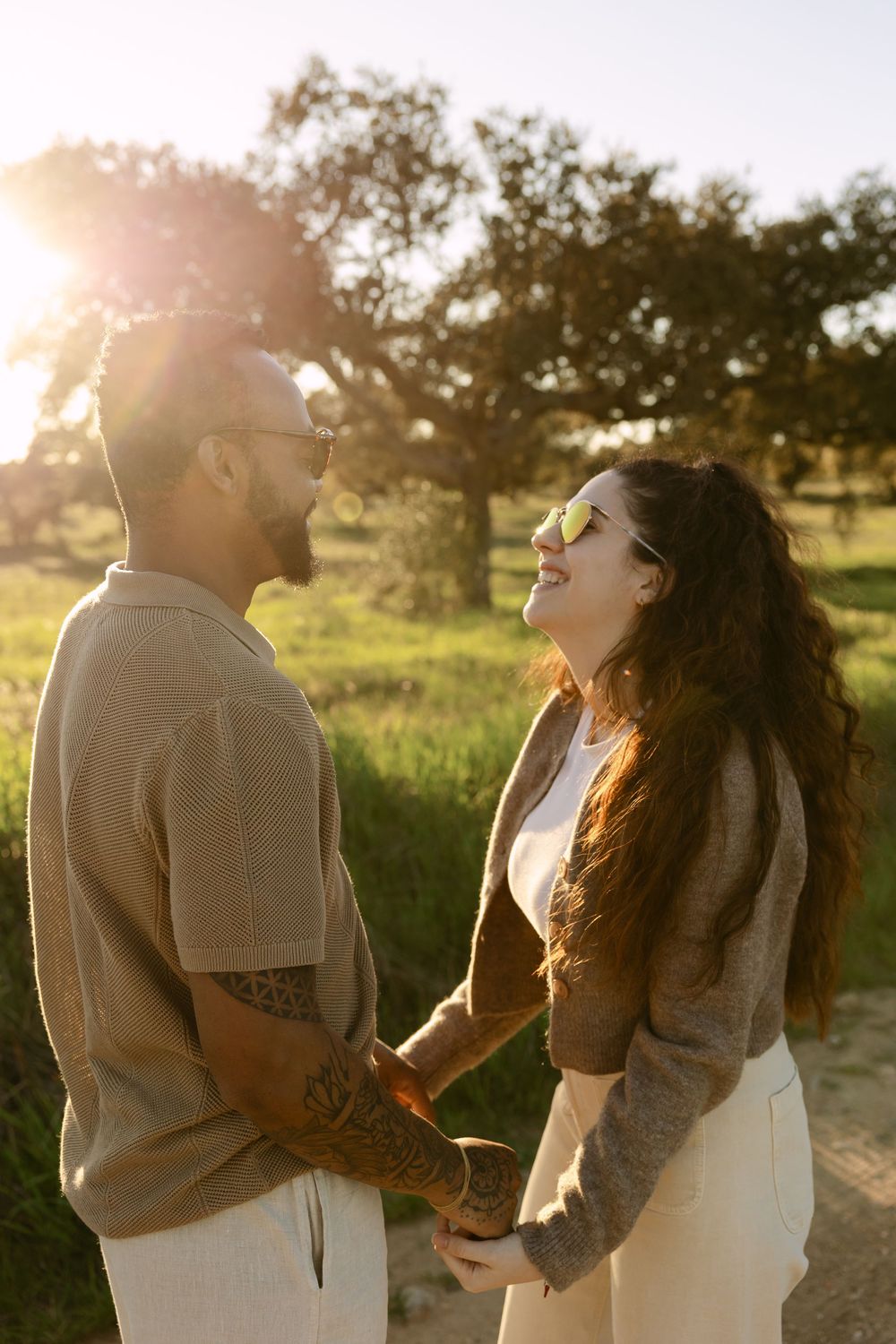 Couple holding hands and smiling at each other in a sunlit park with trees and green grass in the background.