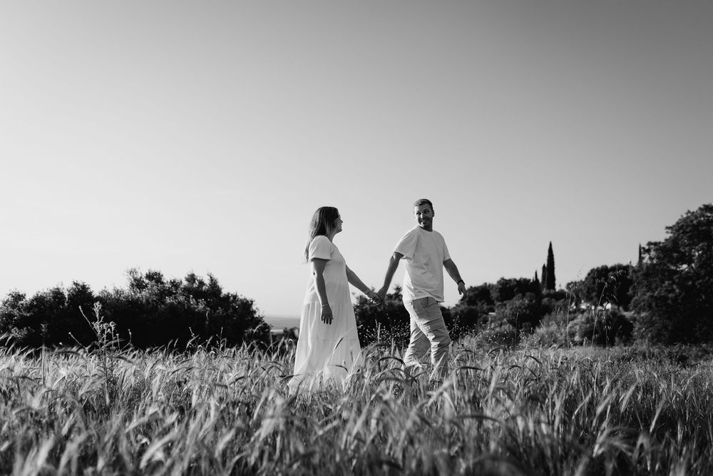 Black and white photo of a couple holding hands walking through a grassy field with trees in the background.