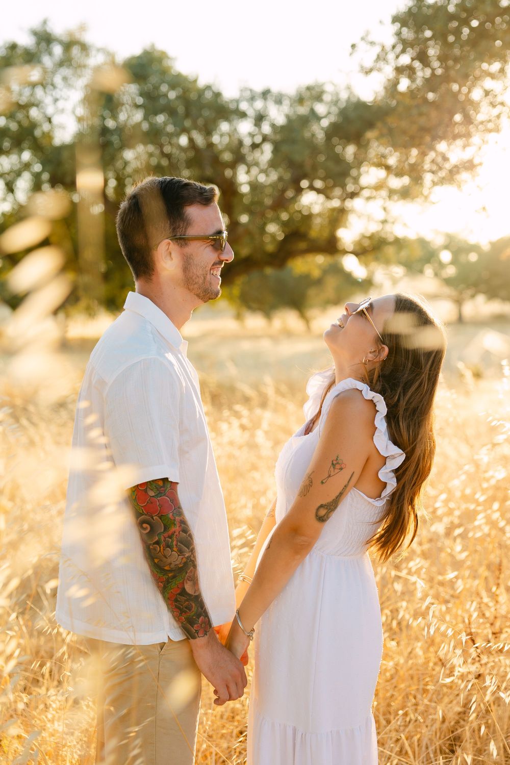 A couple holds hands and laughs together in a golden sunlit field, the man tattooed in a white shirt, woman in a white dress.