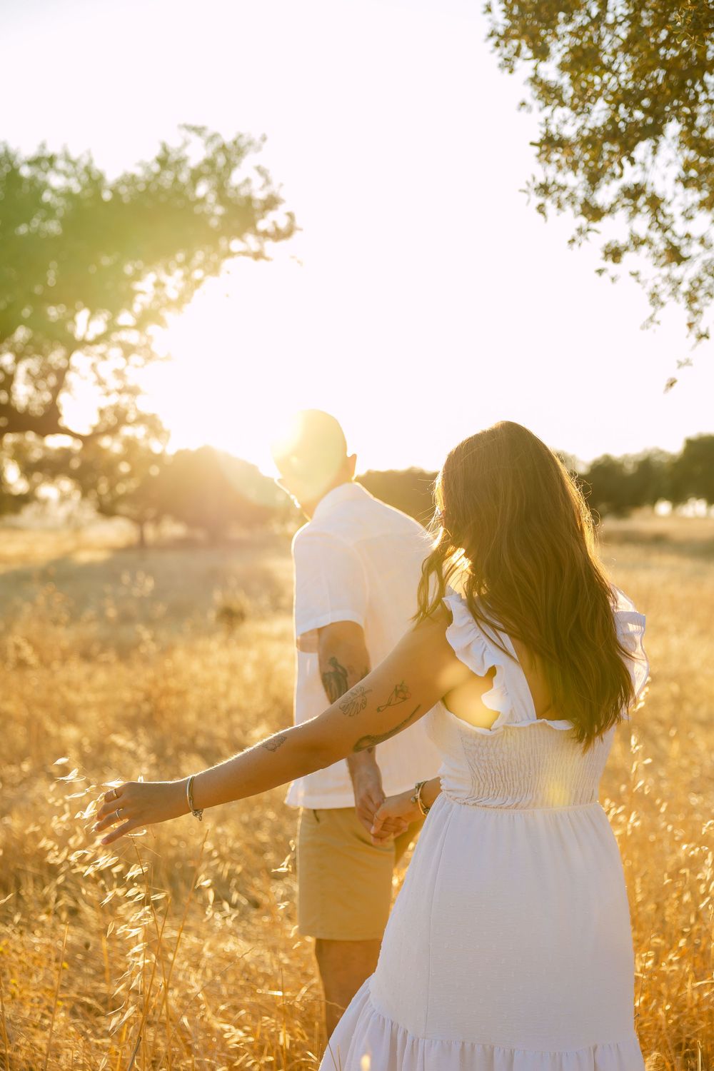 Couple walking hand in hand through golden sunlit field, woman in white dress, man in white shirt, warm sunset glow.