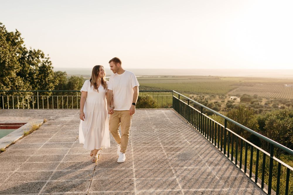 Couple walking hand in hand on a sunny terrace overlooking green fields at golden hour.