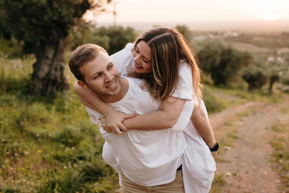 Happy couple sharing a piggyback ride on a scenic dirt path at sunset, both wearing white shirts.