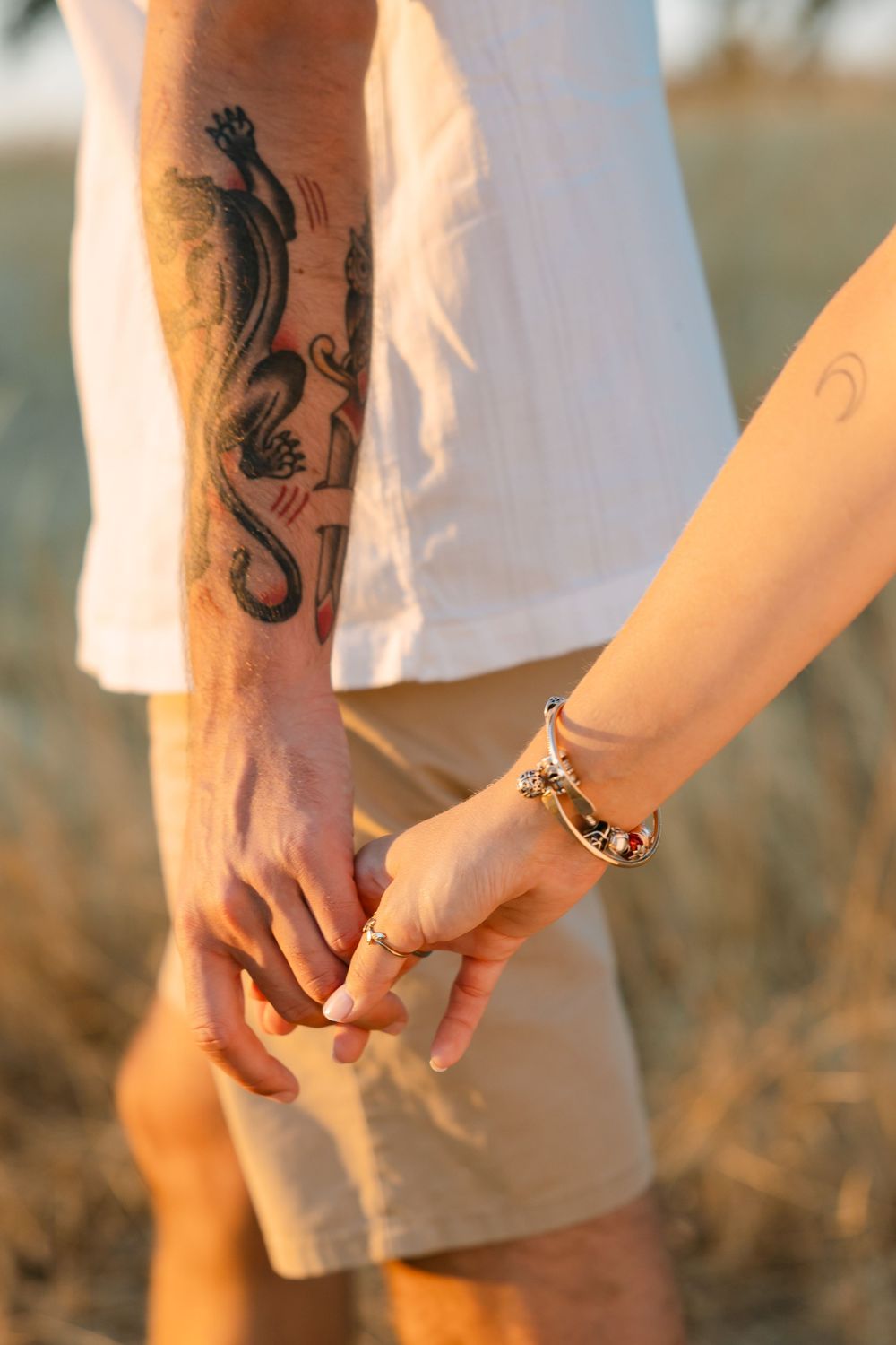 Couple holding hands at sunset, man with dragon tattoo on forearm, woman wearing bracelets, outdoor field background.