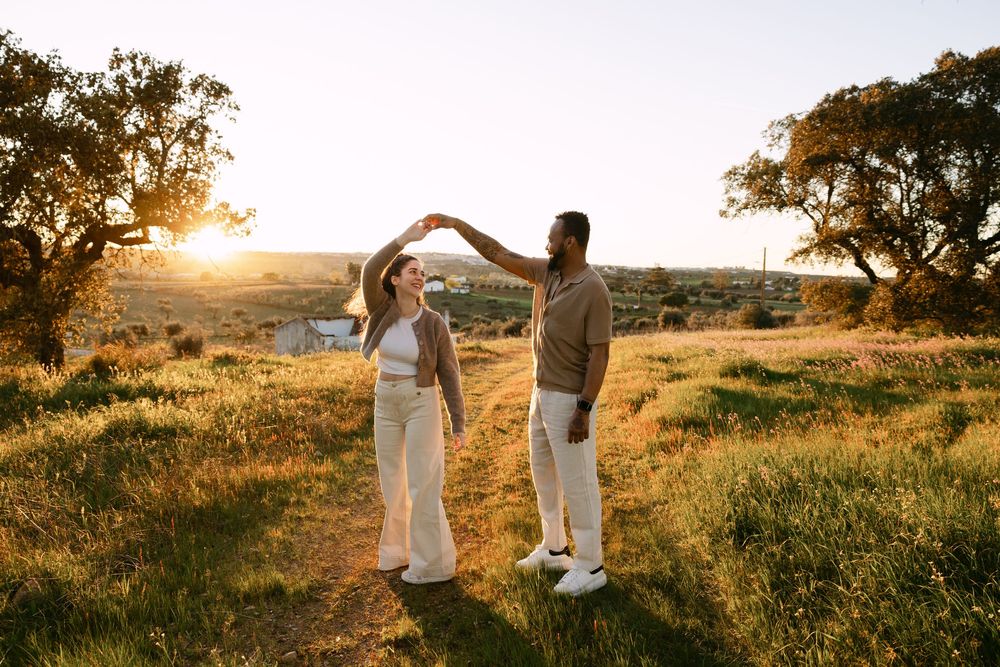Couple dancing at sunset in a golden meadow with oak trees, man spinning woman on a scenic countryside path.