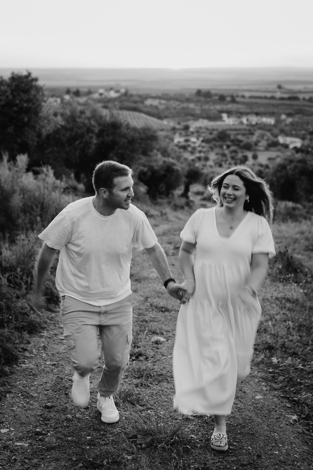 Black and white photo of a happy couple holding hands and walking on a scenic hilltop path surrounded by trees.