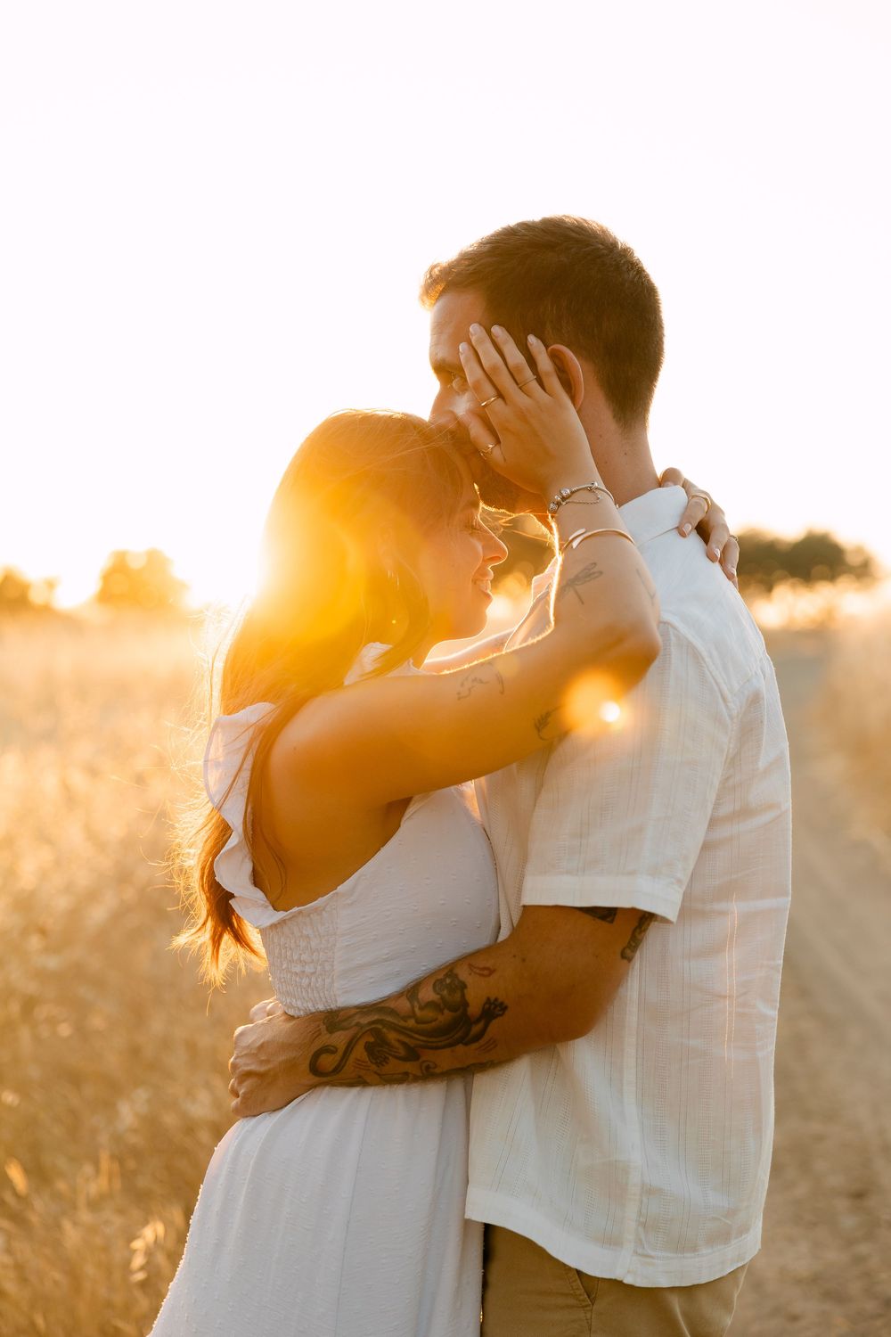 Couple embracing at golden hour in a wheat field, backlit by warm sunset glow, woman in white dress with tattoos.