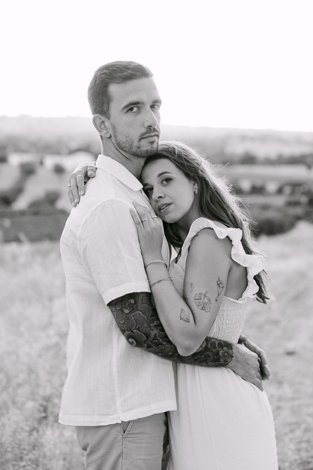 Black and white photo of a couple embracing outdoors, woman in white dress leaning on tattooed man in white shirt.