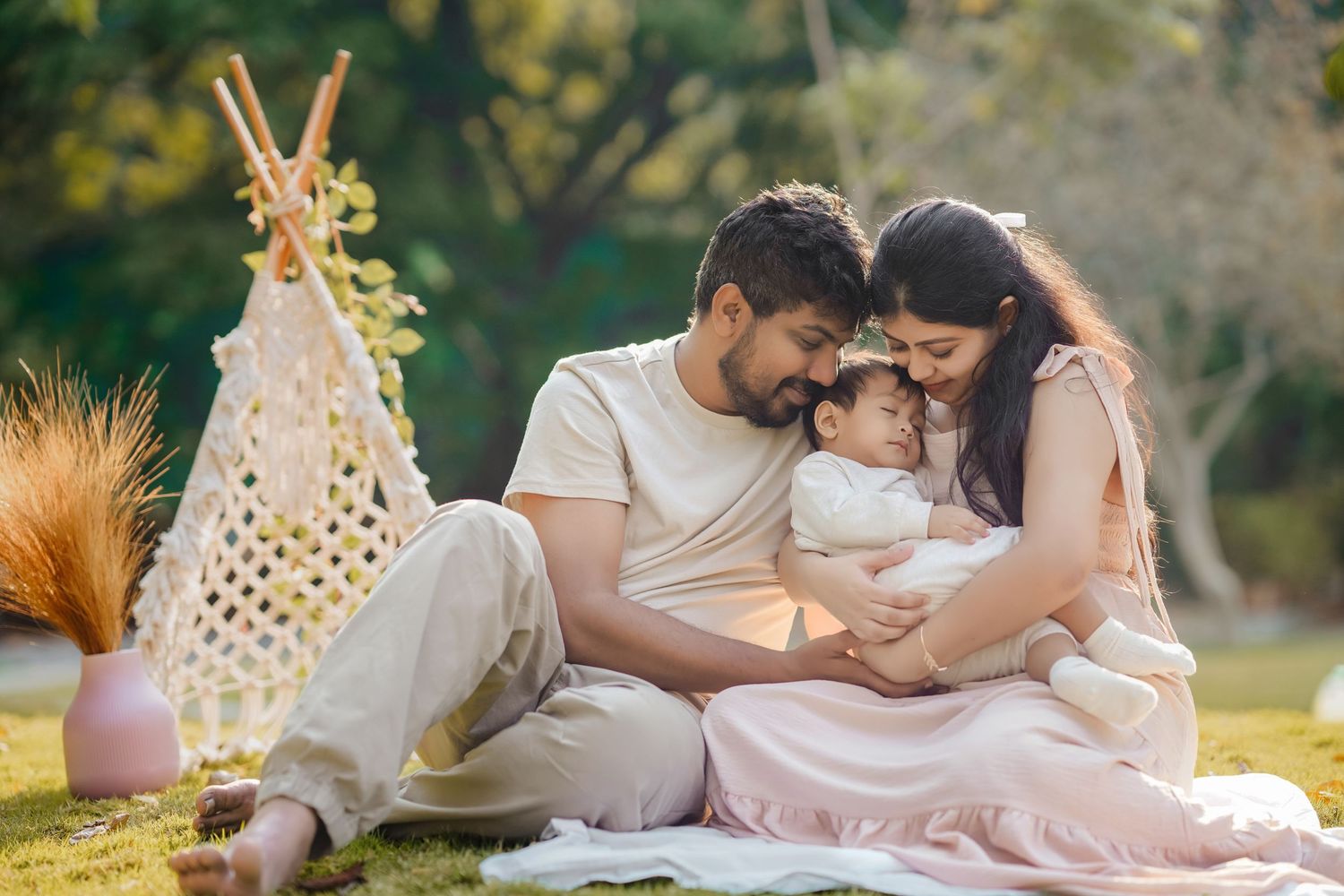 A young Indian family sits outdoors on a blanket, parents lovingly gazing at their newborn baby near a boho teepee.