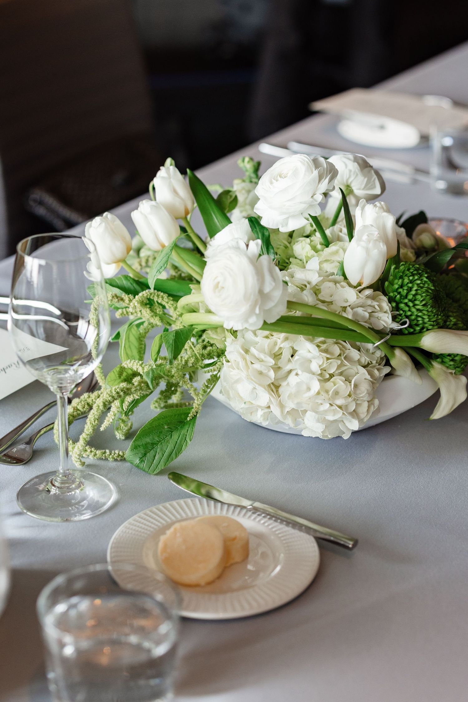 Elegant white floral centerpiece with tulips and hydrangeas on a formal dining table with glassware and a small cookie.