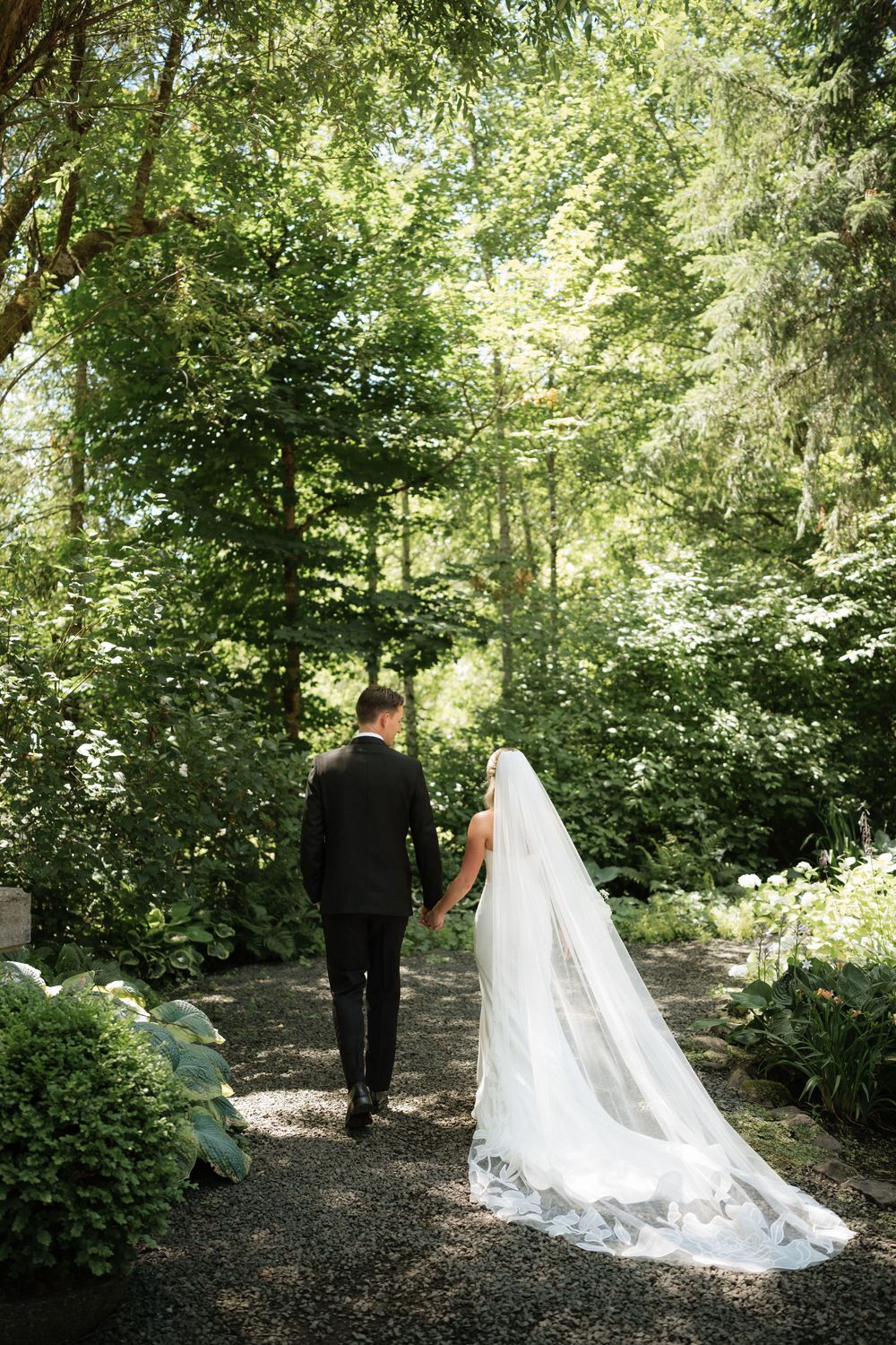 Bride and groom walk hand-in-hand through a lush green garden path, bride wearing a long flowing veil.