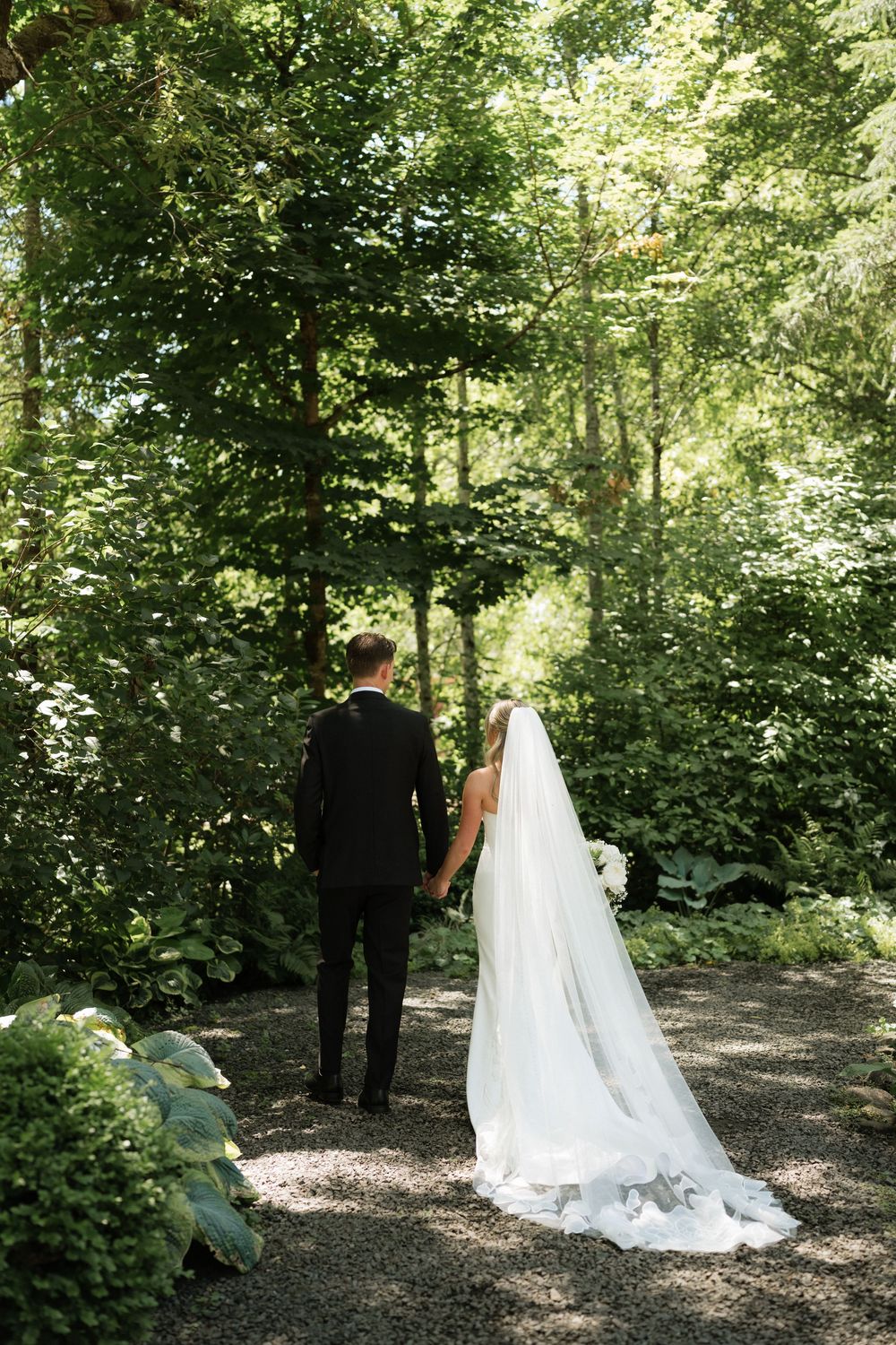 Bride and groom holding hands walking down a garden path surrounded by lush green trees on their wedding day.