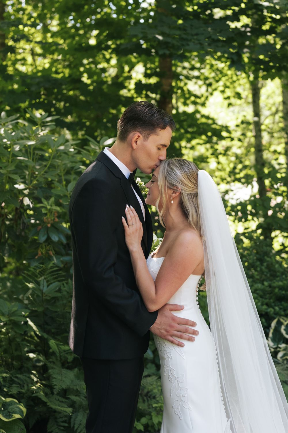 Bride and groom share a romantic moment outdoors, surrounded by lush green trees, as groom kisses bride's forehead.
