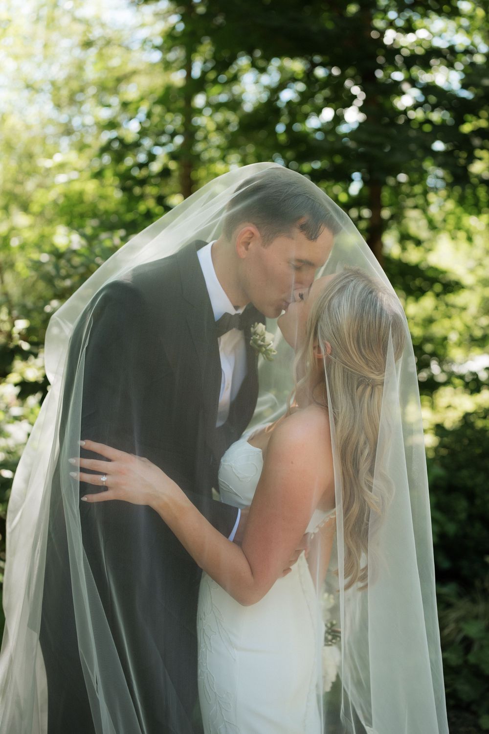 Bride and groom kissing under a flowing veil outdoors, surrounded by lush green trees on their wedding day.