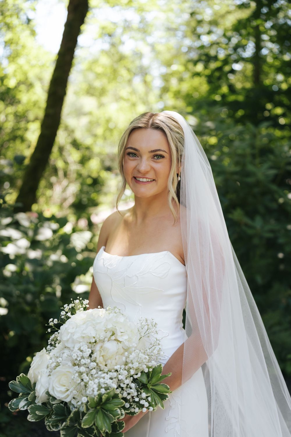 Smiling bride in strapless white gown and veil holding white floral bouquet outdoors among green trees.
