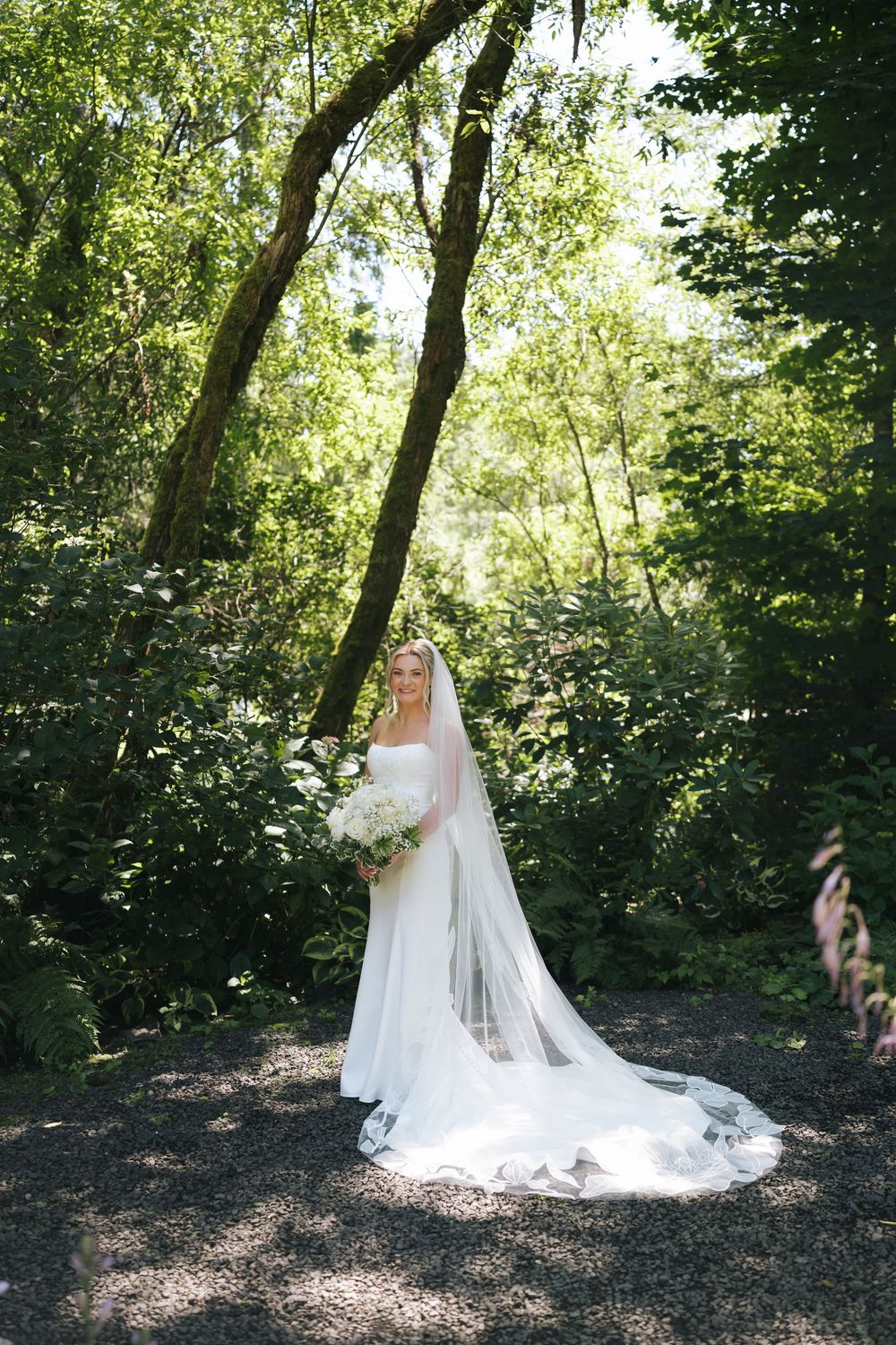 Bride in white gown and veil holds bouquet in lush green forest setting.