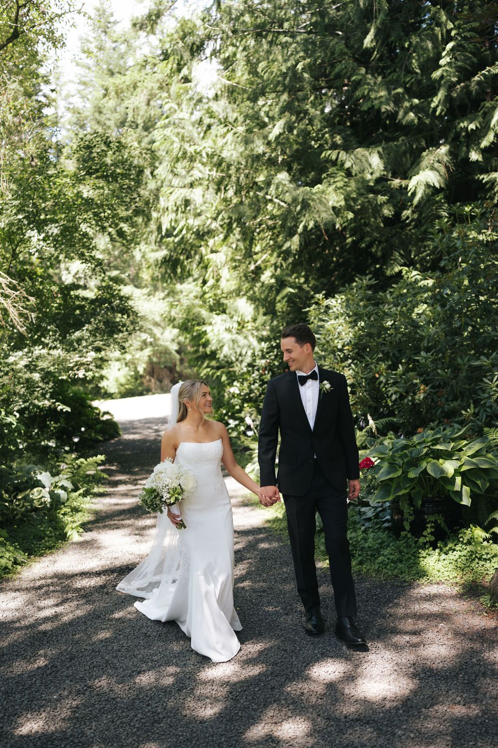 Bride and groom walk hand-in-hand on a tree-lined path, she in a white gown, he in a black tuxedo.