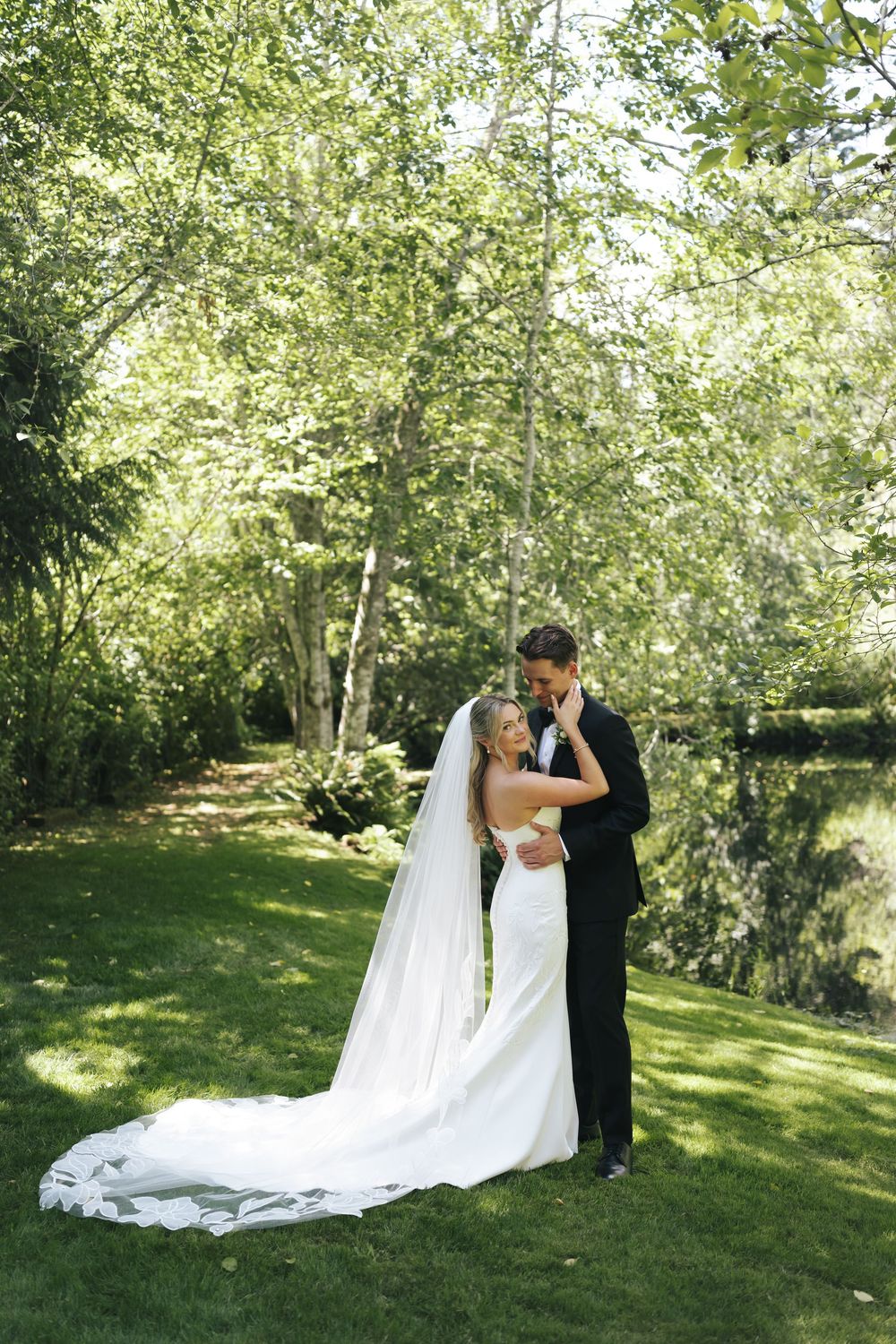 Bride and groom embrace on green lawn surrounded by trees on wedding day.