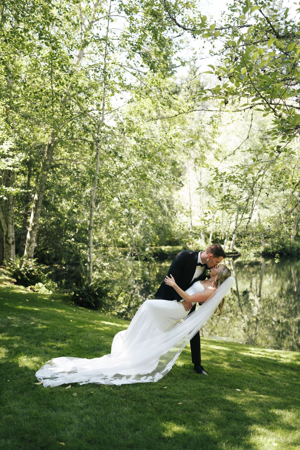 Groom dips bride in white gown for romantic kiss on lush green lawn near reflective pond.