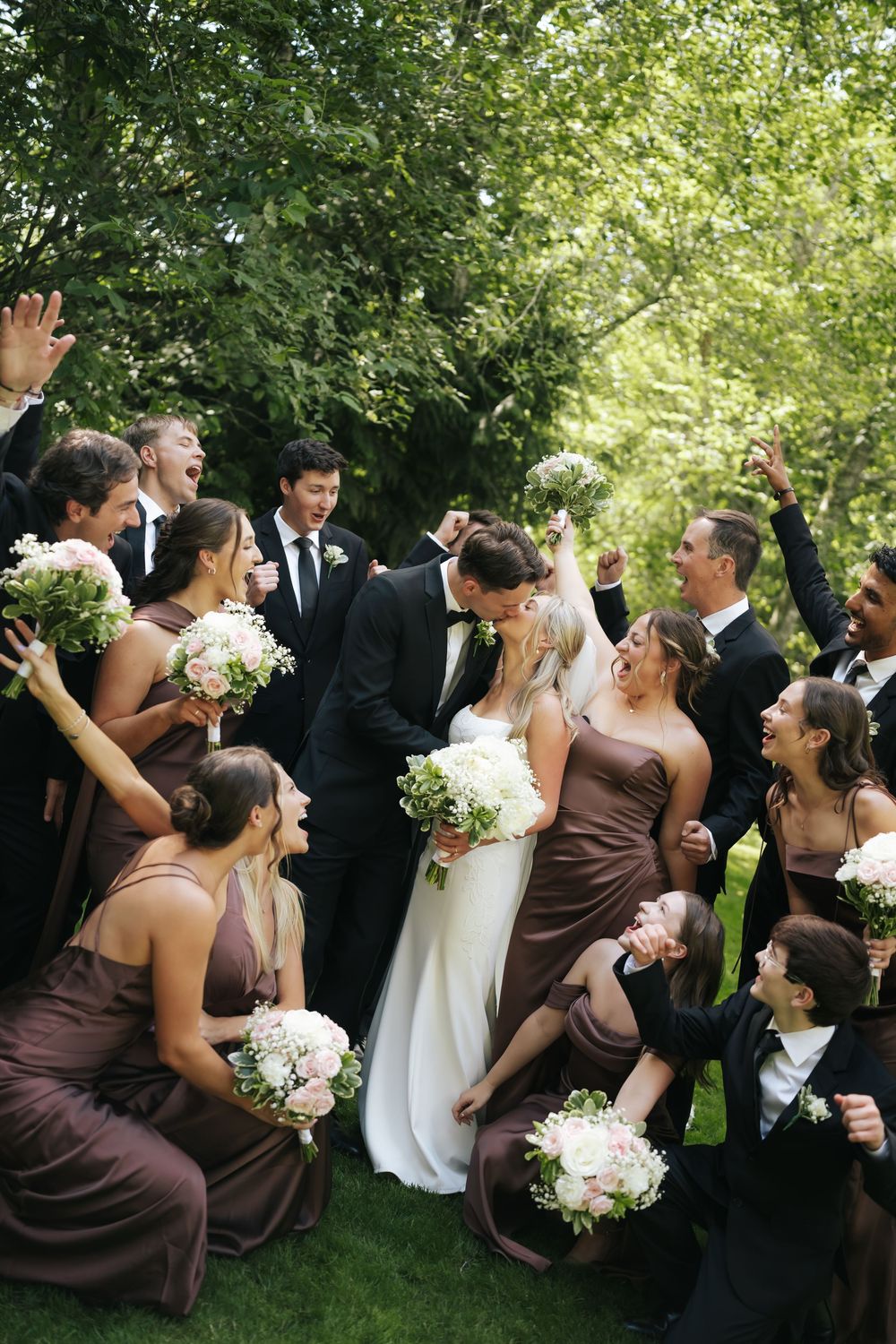 Wedding party celebrates as bride and groom kiss, surrounded by bridesmaids in brown dresses and groomsmen in black suits.