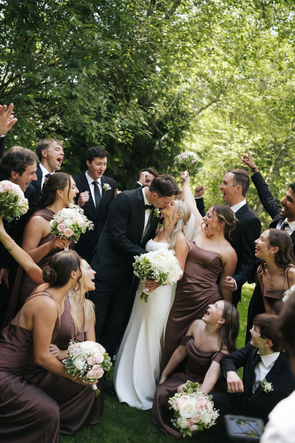 Bride and groom share a kiss surrounded by cheering wedding party in brown bridesmaid dresses.
