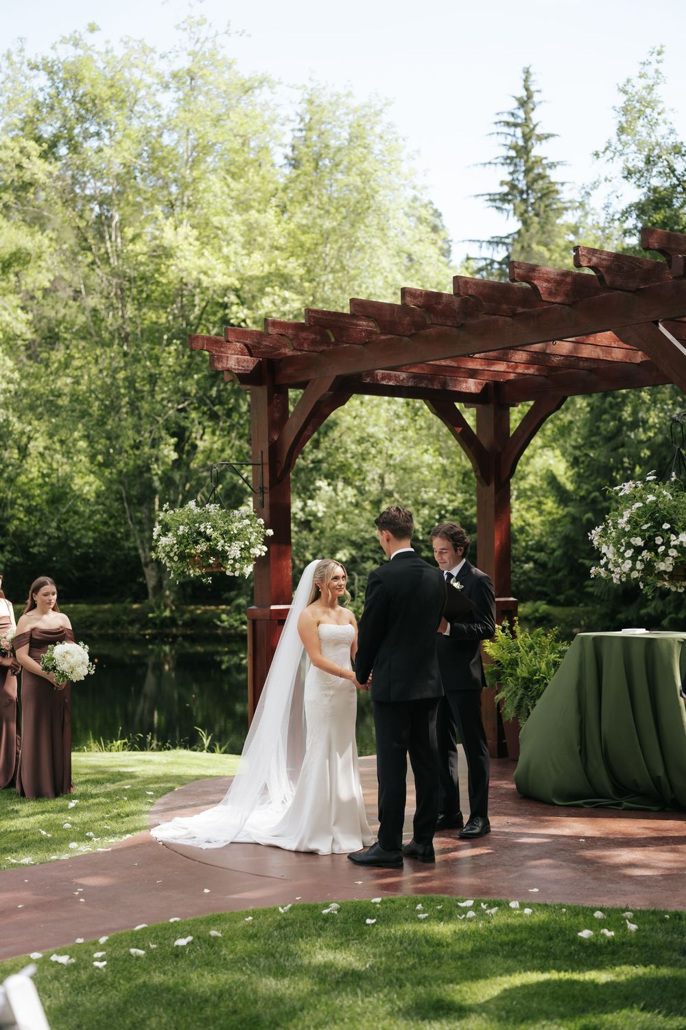 Bride and groom exchange vows under a rustic wooden pergola surrounded by lush greenery at an outdoor garden wedding ceremony.