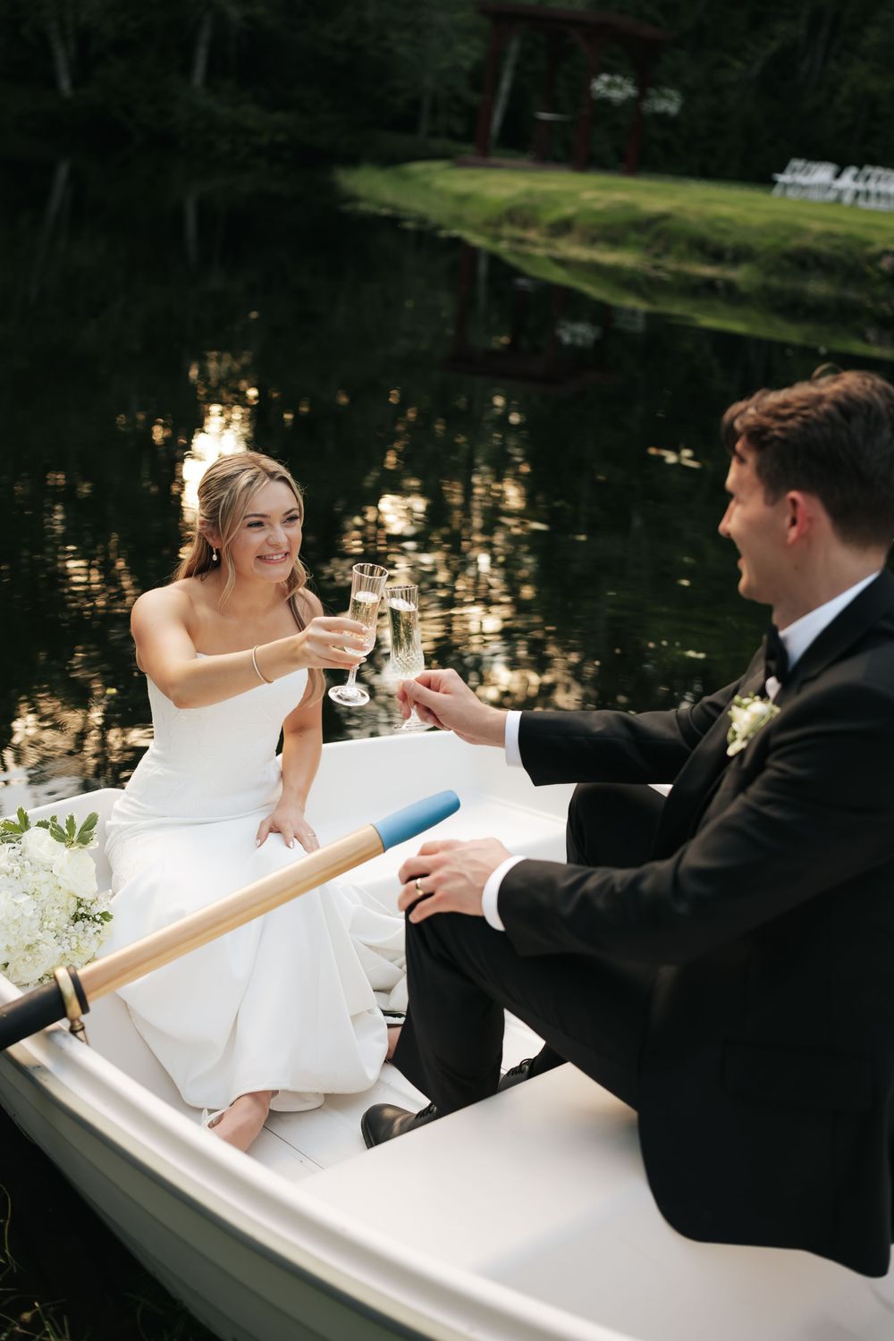 Bride and groom toasting champagne glasses while sitting in a rowboat on a serene lake on their wedding day.