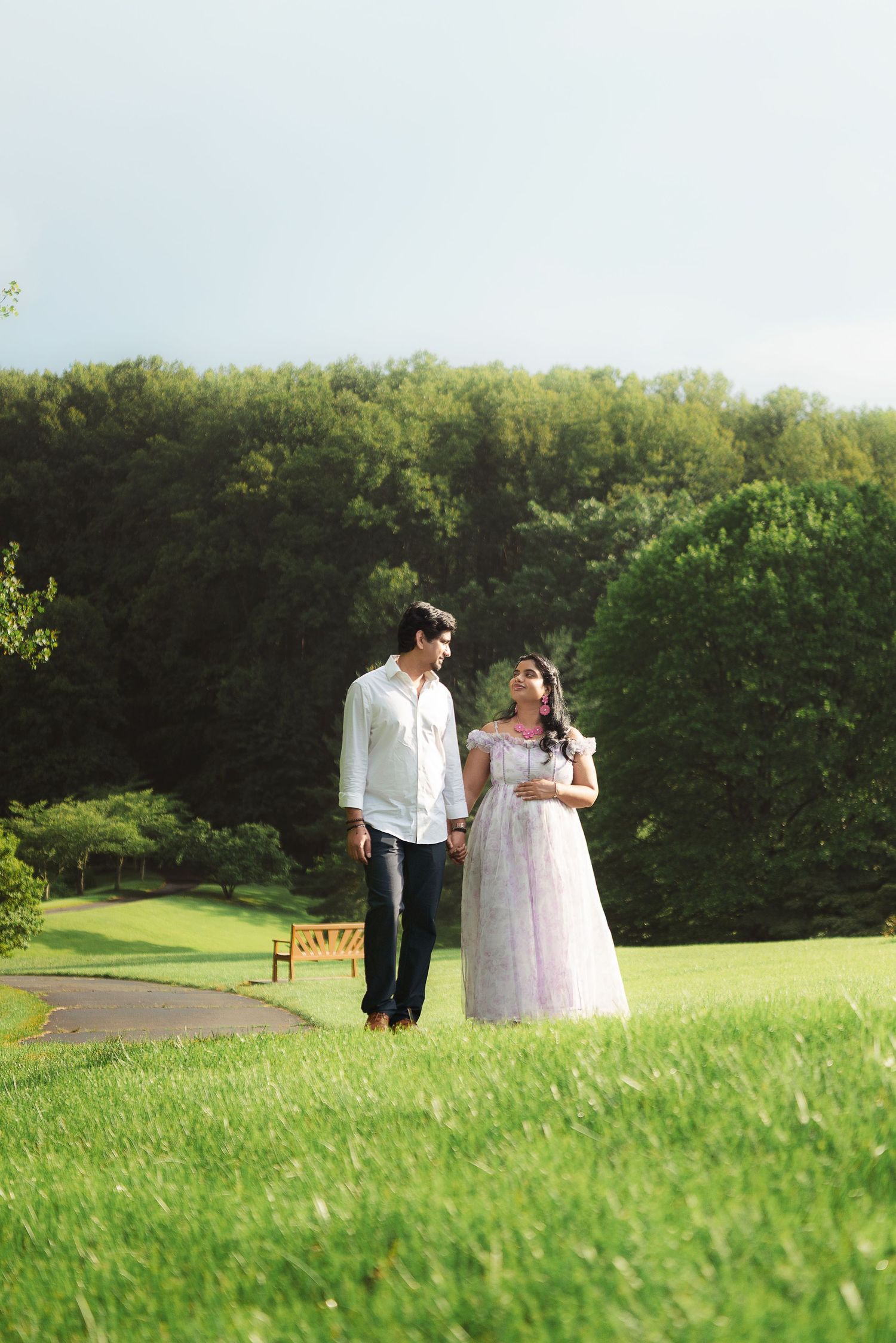 A couple walks hand-in-hand on lush green grass, surrounded by trees, the woman in a white gown and man in a white shirt.