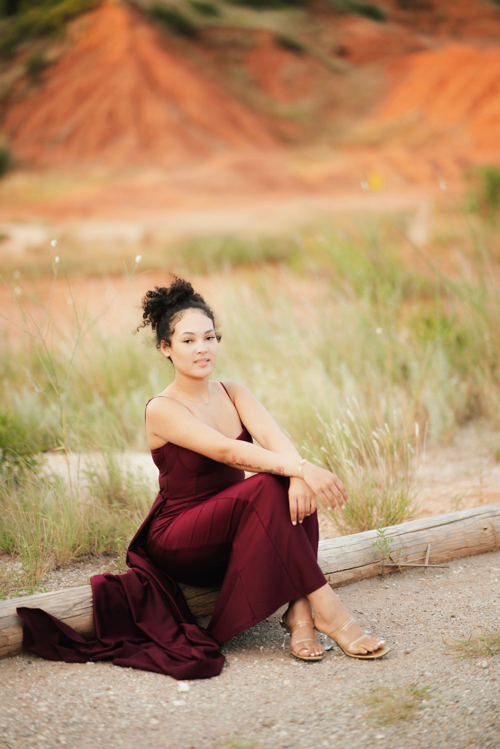 Young woman in burgundy dress sitting on log in sandy field with red clay hills backdrop.
