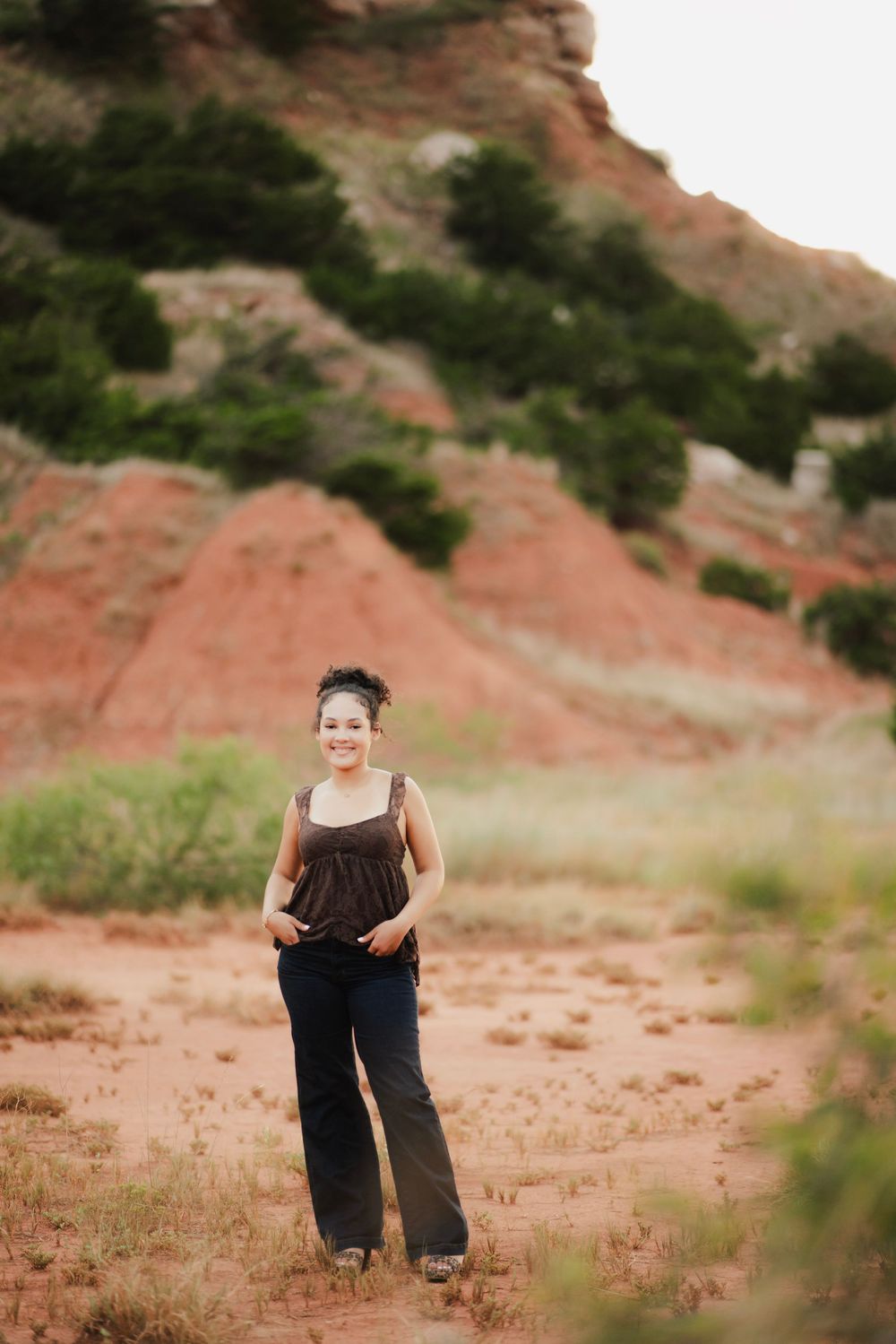 Smiling woman in brown top and jeans standing on red dirt trail with rocky hills behind her.