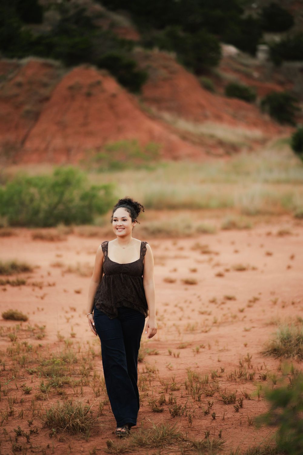 Smiling woman in black top and jeans walking on red sand with eroded clay hills in background.
