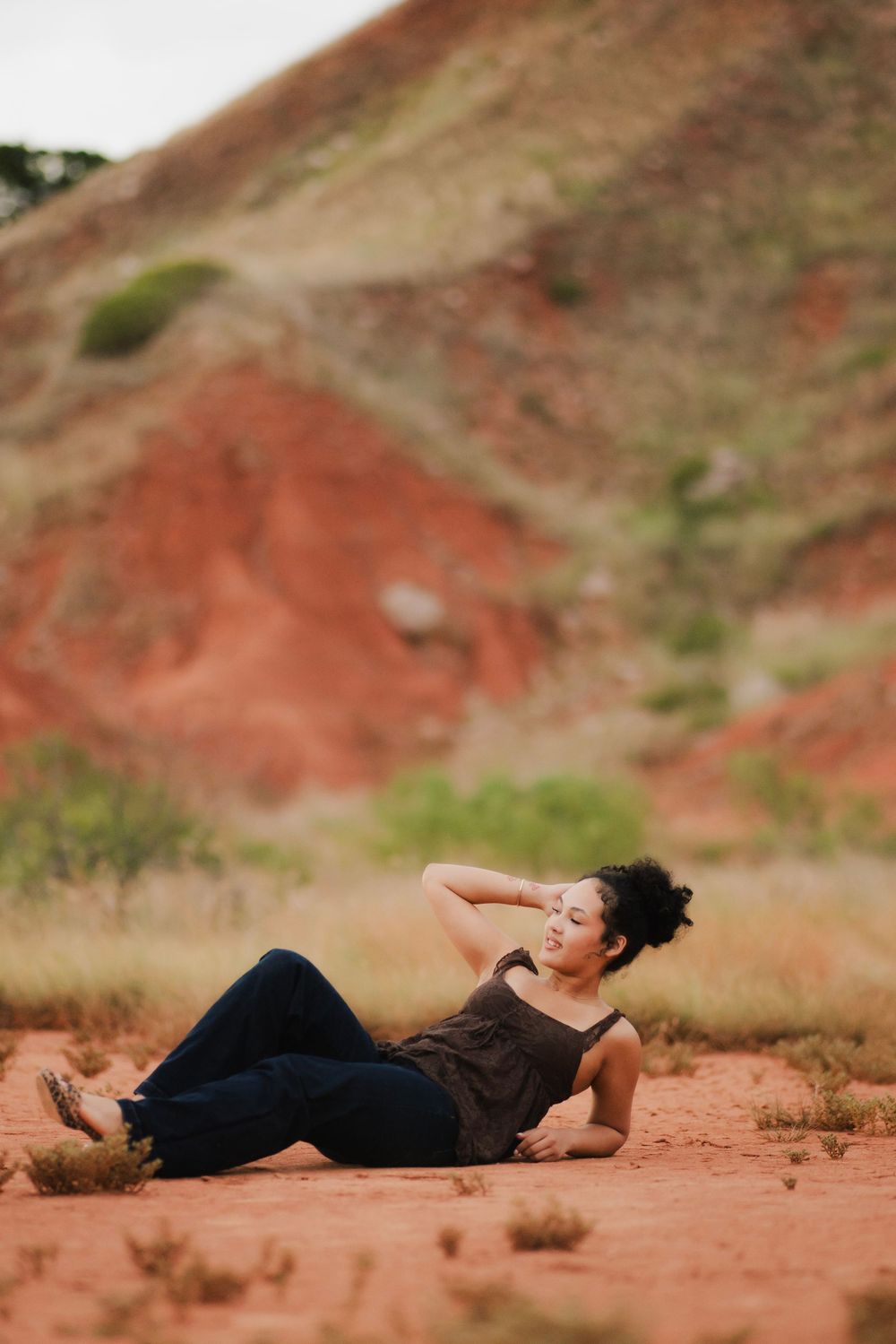 Woman in jeans and tank top reclining on red desert dirt with rocky hillside background.