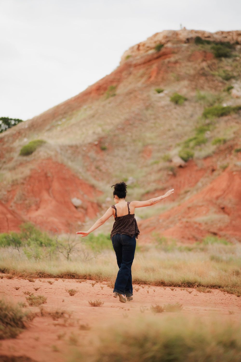 Woman in brown top and jeans walking away with arms outstretched toward red clay hillside.