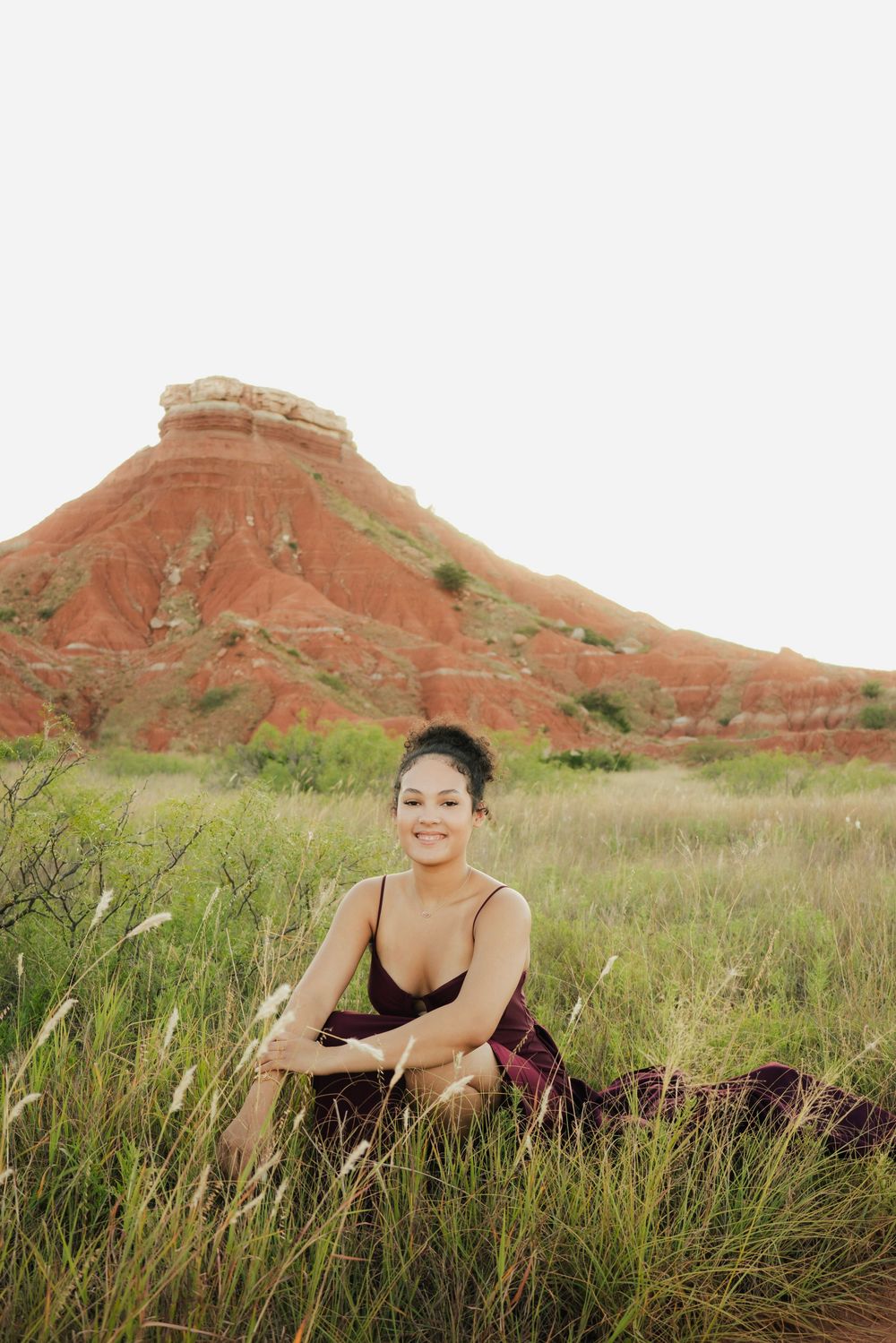 Woman in burgundy dress sits smiling in tall grass with red rock butte formation in background.