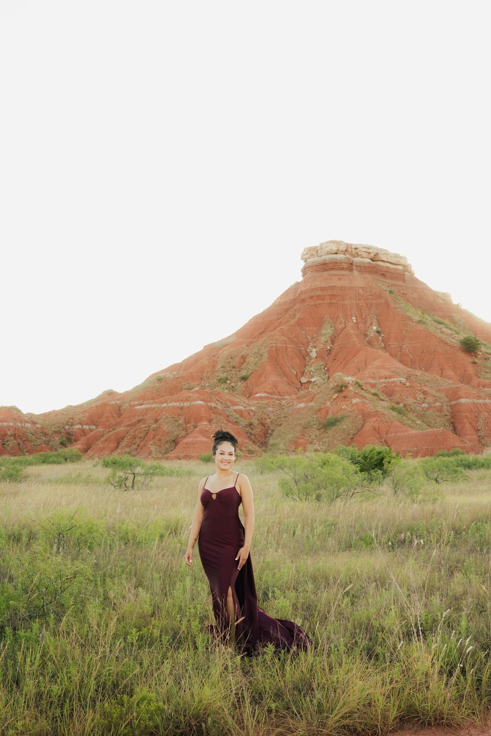 Woman in burgundy gown stands in grassy field with red rock butte formation in background.