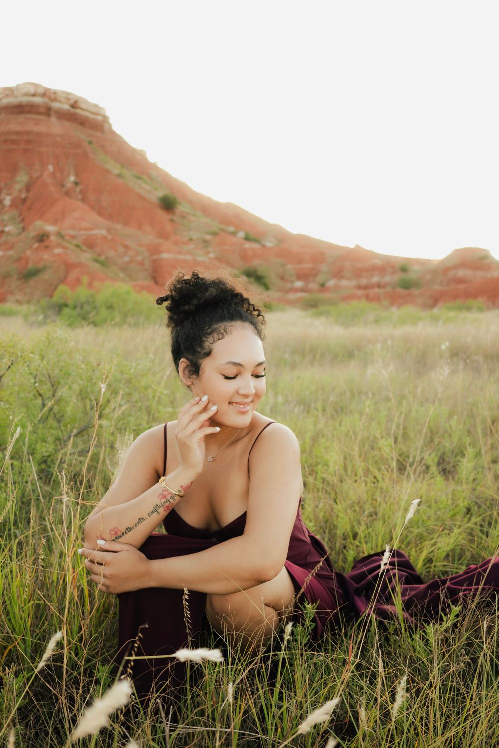 Woman in burgundy dress sitting in green field with red rock formations in background.