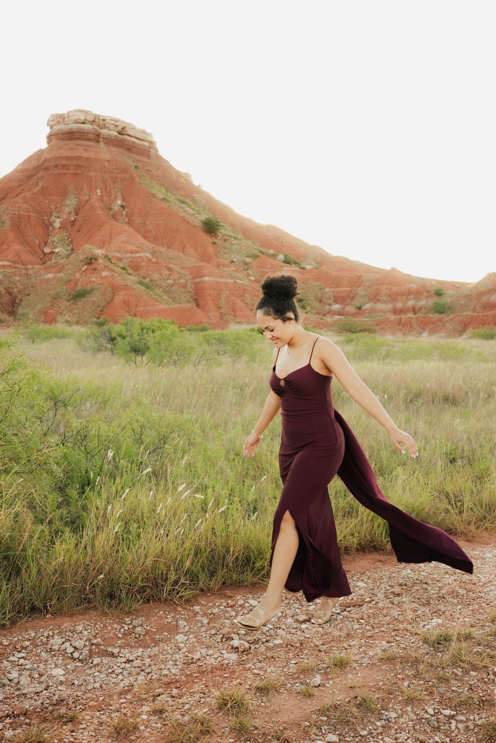 Woman in burgundy gown walks through red rock canyon landscape with lush green grass and sandstone formations.