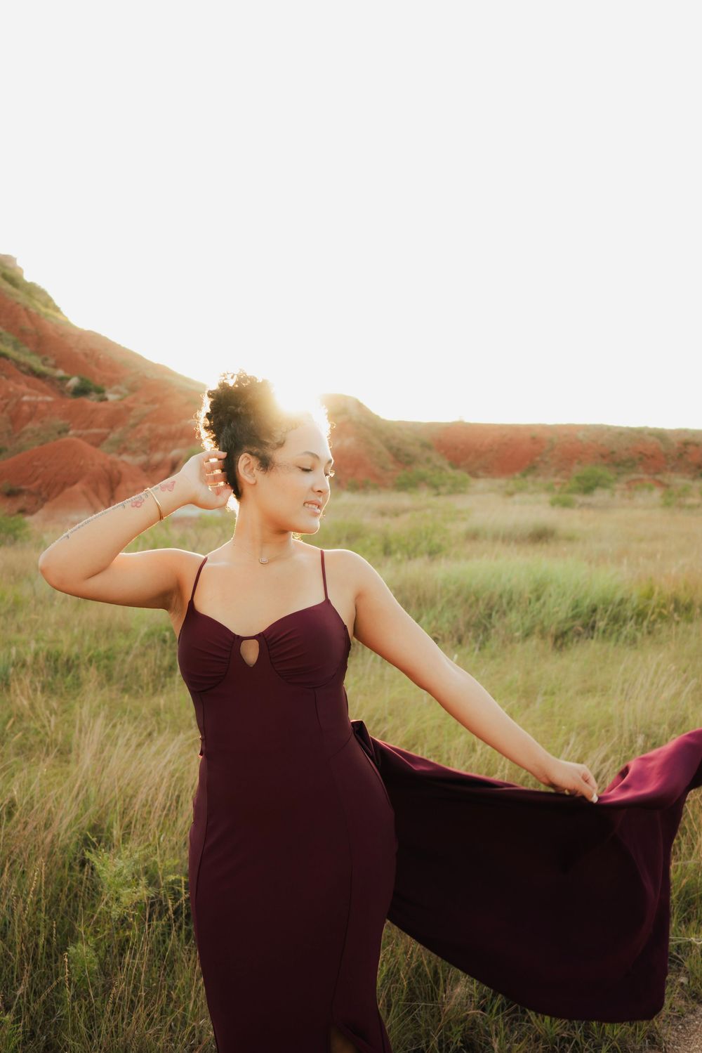 Woman in burgundy gown twirling in golden hour sunlight against red rock hills and green meadow.