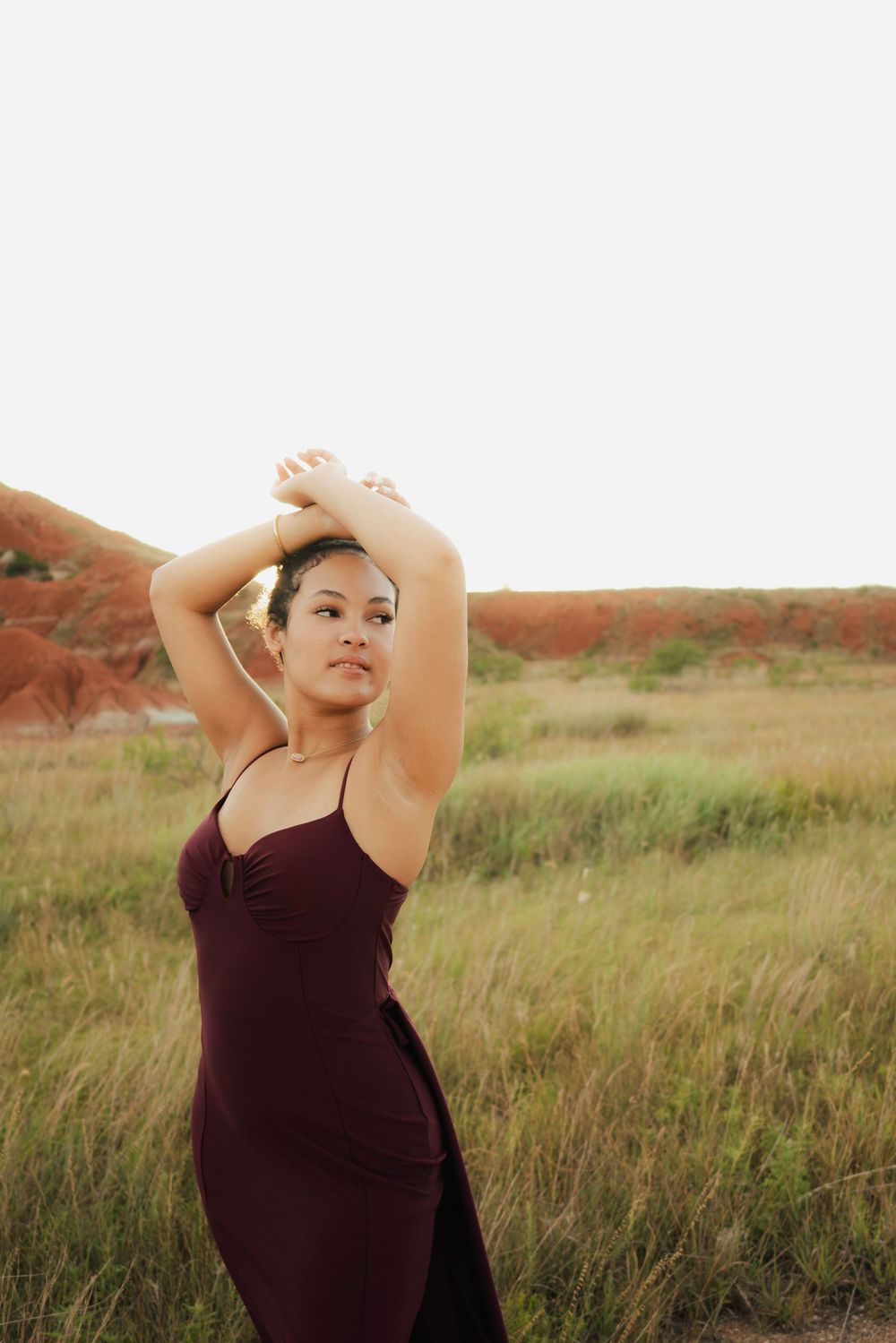 Woman in burgundy dress posing with arms raised in a grassy field with red rock formations in background.