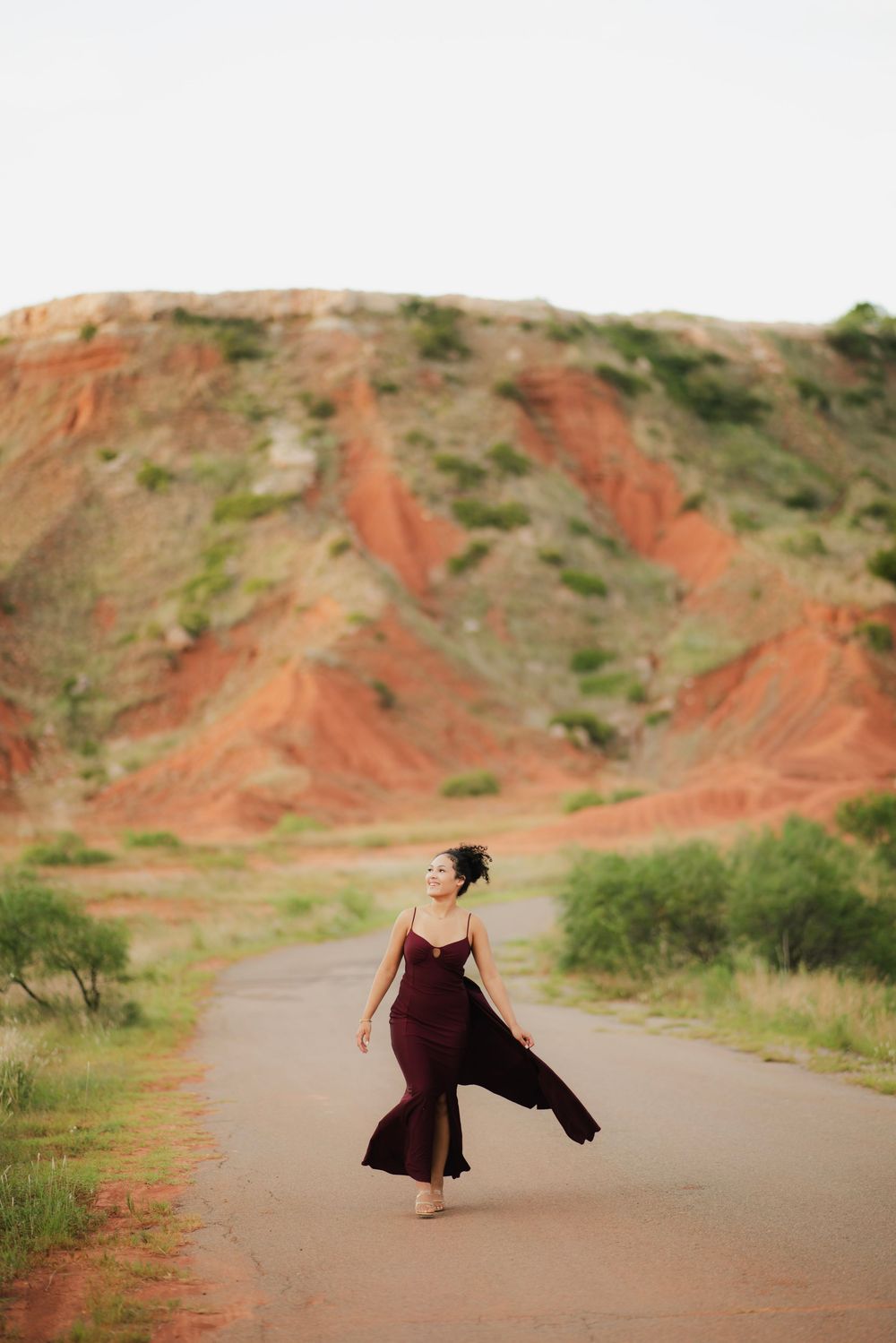 Woman in flowing burgundy dress walking on road with red rock formations in background.