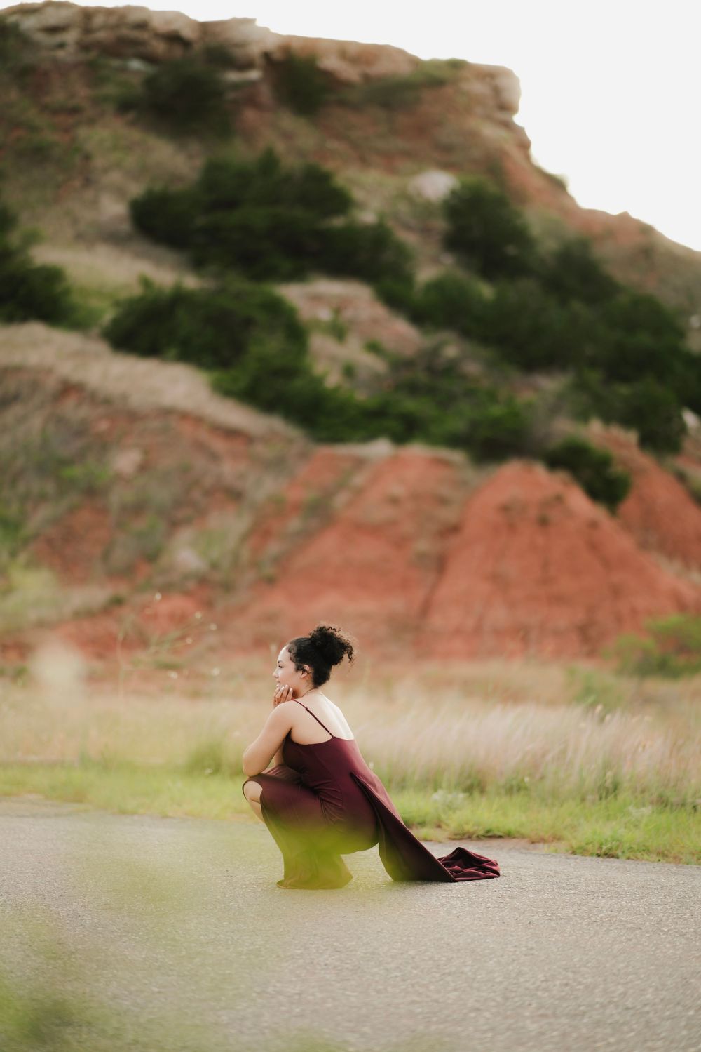 Woman in burgundy gown sits gracefully on road with red clay hills and greenery behind her.