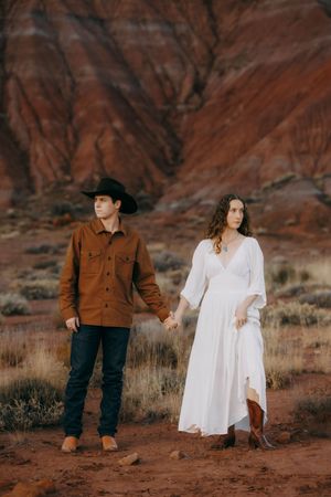 Couple holding hands in red rock desert, man in cowboy hat and brown jacket, woman in white boho dress and boots.