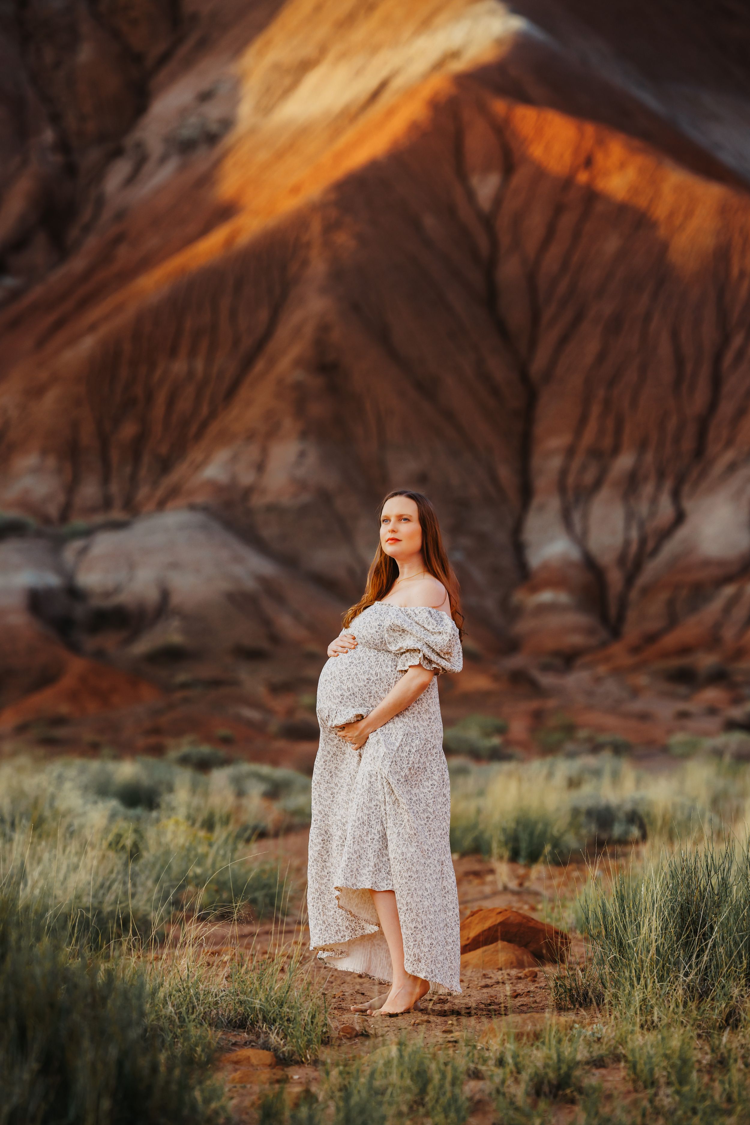 Pregnant woman in white floral dress standing in desert landscape with red rock formations at golden hour.