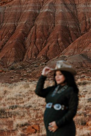 Pregnant woman in black dress and cowboy hat poses before dramatic red rock badlands formations.