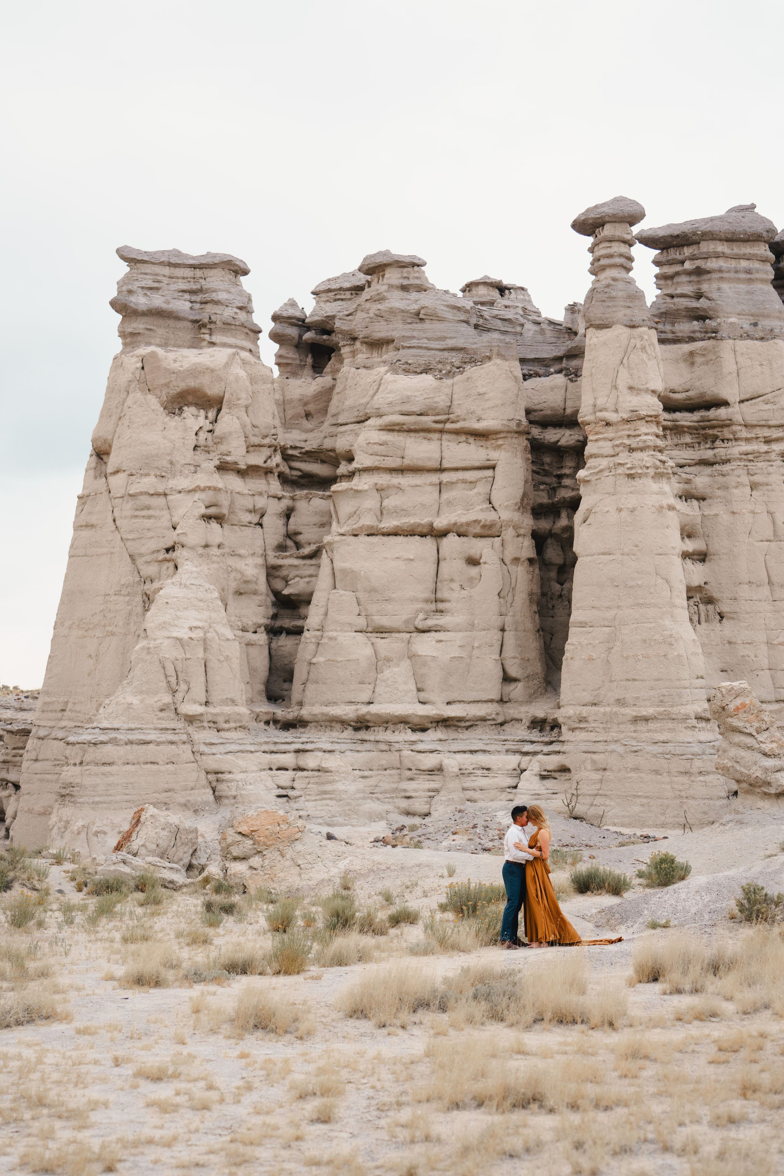 Couple embracing before towering white sandstone hoodoo formations in a dramatic desert landscape.