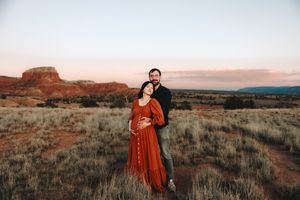 Couple embracing in red desert landscape at sunset, pregnant woman in rust dress, mesa formations in background.