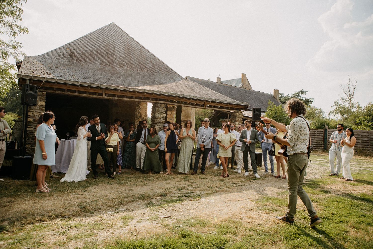 Outdoor wedding gathering with guests assembled before a rustic barn venue on a sunny day.