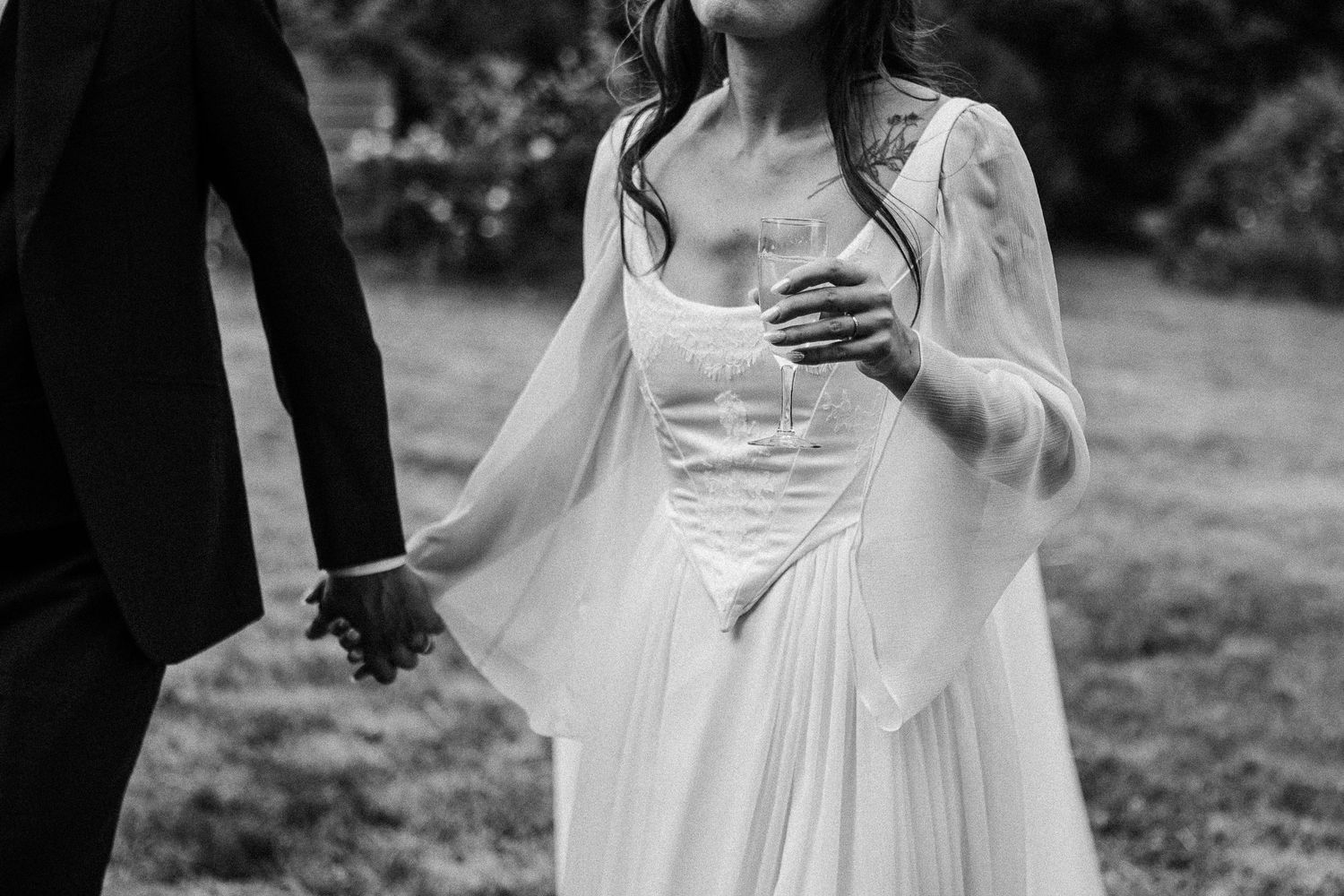 Black and white photo of a bride in a flowing white gown holding hands with a groom outdoors.
