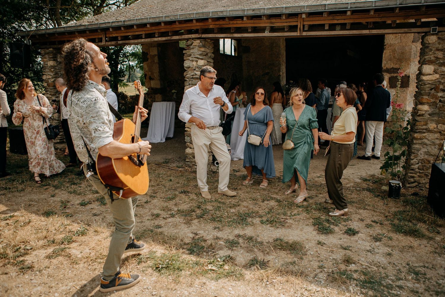 Guitarist performs outdoors at a rustic barn wedding reception while guests dance and celebrate in summer.