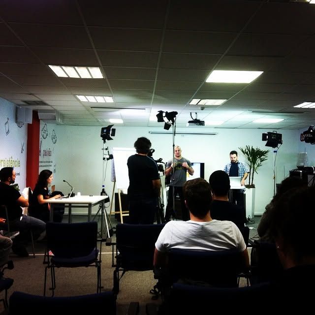 A film crew records a speaker at an indoor seminar with audience members seated in chairs under bright studio lights.