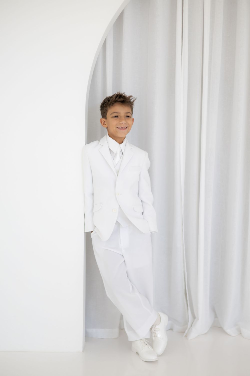 A smiling boy in a white suit, vest, and tie poses confidently against a white wall with curtains.